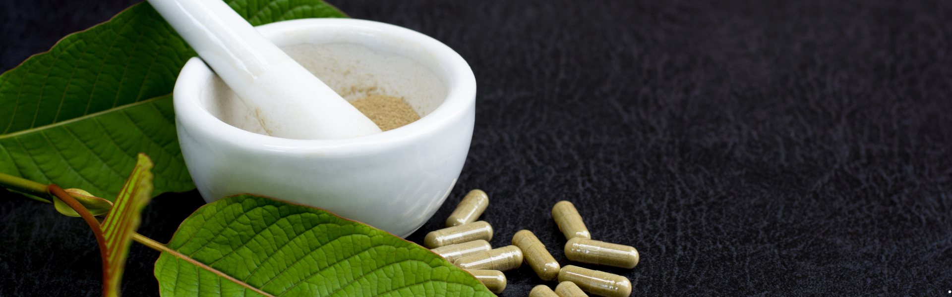 kratom leaves sitting next to a mortar and pestle with some small capsules next to them
