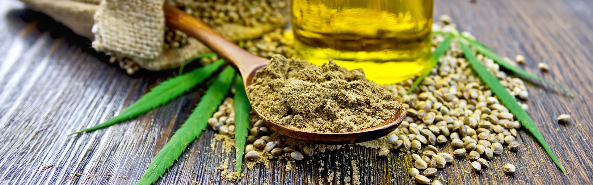 a wooden spoon holding ground marijuana lays on marijuana seeds and a leaf next to a bottle of oil on a wooden table