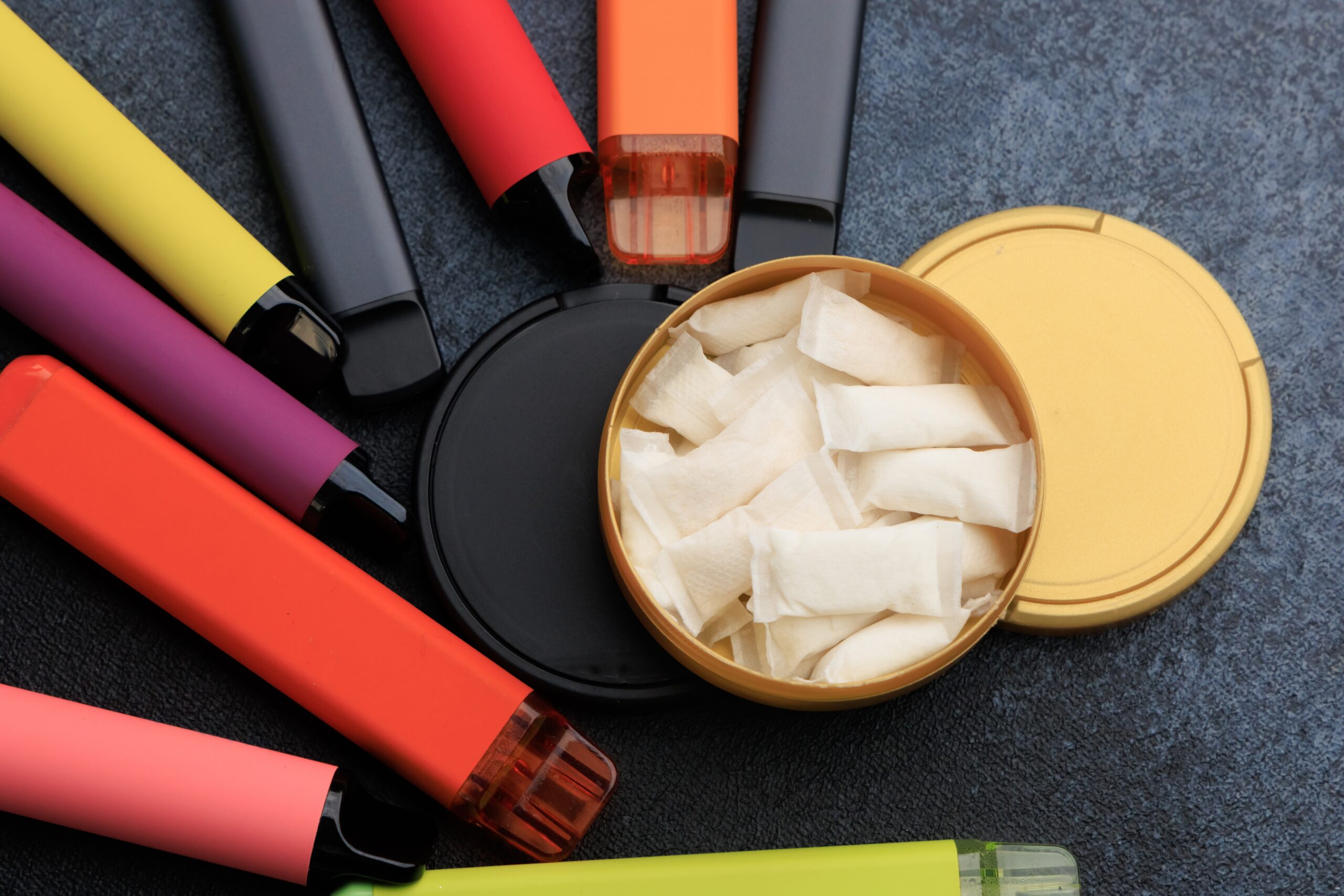 A round container filled with white nicotine pouches next to several colorful disposable vape devices.