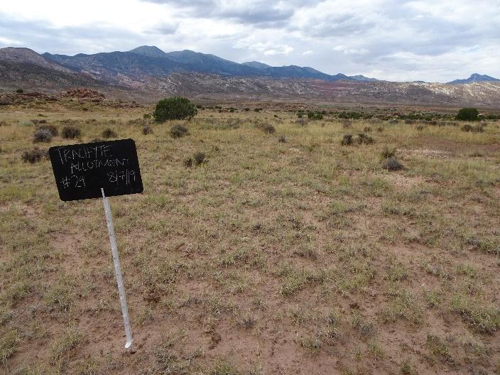 a sandy ground with grasses poking through and a black sign that reads TRACAYTE ALLOTMENT #24 8/7/19 in the foreground. The midground shows some higher bushes and trees. the background is larger hills and mountains.